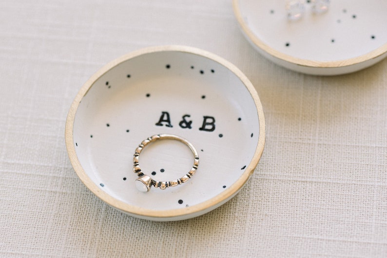 May include: A white ceramic ring dish with black polka dots and the text "A&B." A gold ring with a white stone is in the dish.