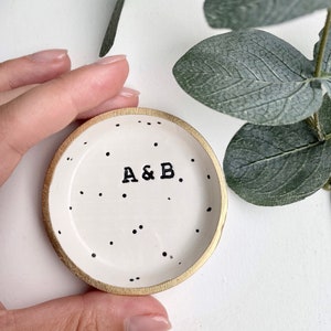 May include: A white ceramic ring dish with a gold rim and black polka dots. The dish has the initials "A & B" written in black.