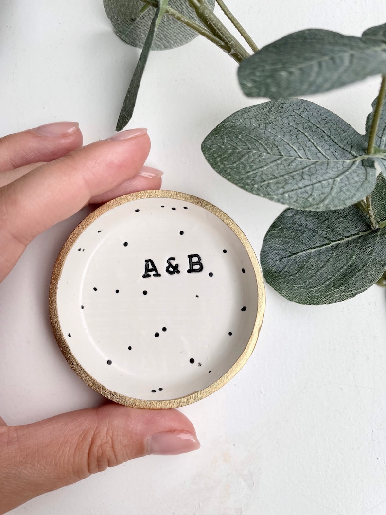 May include: A white ceramic ring dish with a gold rim and black polka dots. The dish has the initials "A & B" written in black.