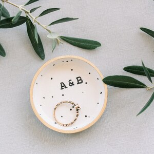 May include: A white ceramic ring dish with black polka dots and the text "A&B." A gold ring is inside the dish.