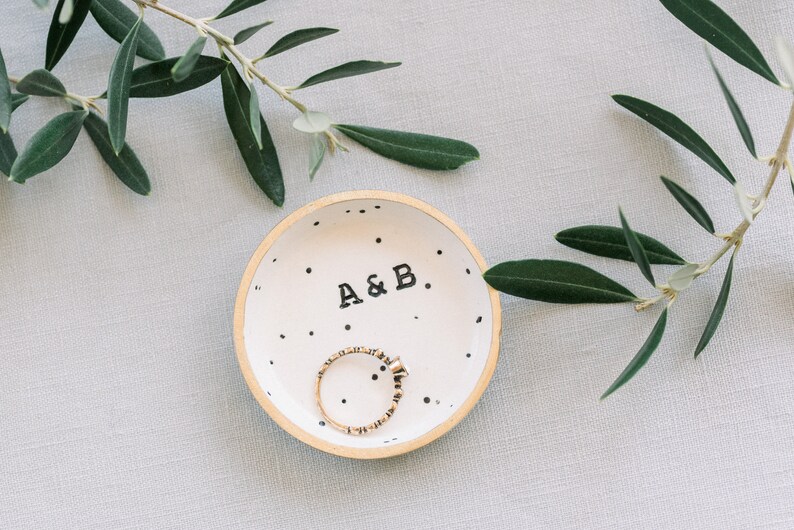 May include: A white ceramic ring dish with black polka dots and the text "A&B." A gold ring is inside the dish.