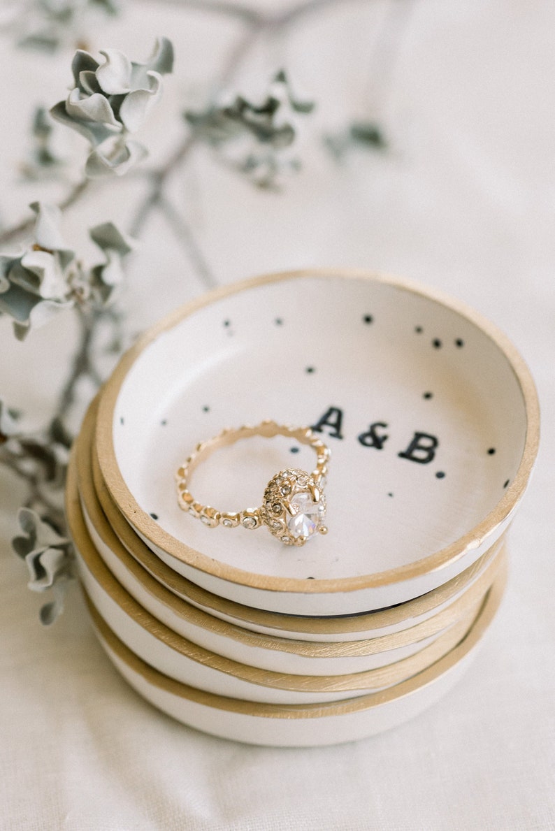May include: A gold ring with a large, clear diamond sits in a white ceramic ring dish with black polka dots and the inscription "A & B". The dish is stacked on top of three other similar dishes.