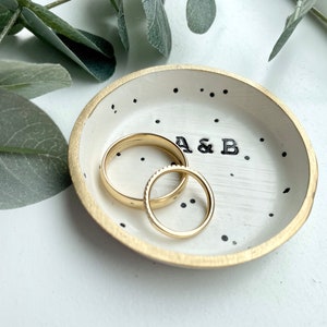 May include: White ceramic ring dish with black polka dots and gold trim. The dish has the initials "A & B" written in black. Two gold wedding bands are sitting in the dish.