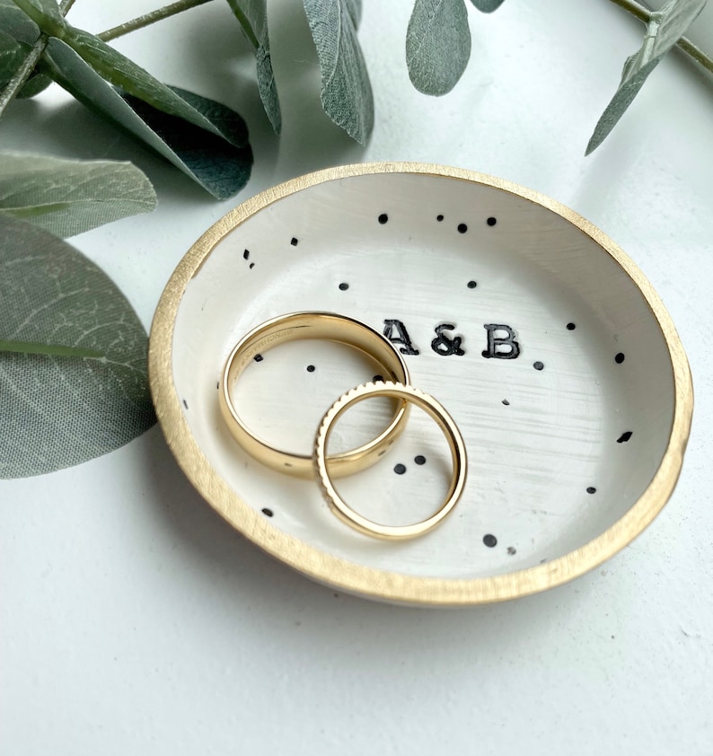 May include: White ceramic ring dish with black polka dots and gold trim. The dish has the initials "A & B" written in black. Two gold wedding bands are sitting in the dish.