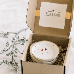 May include: A set of four small white ceramic dishes with black polka dots and a red heart with a black letter K in the center. The dishes are stacked inside a brown cardboard box with a white label that says "LULU & BUNi".