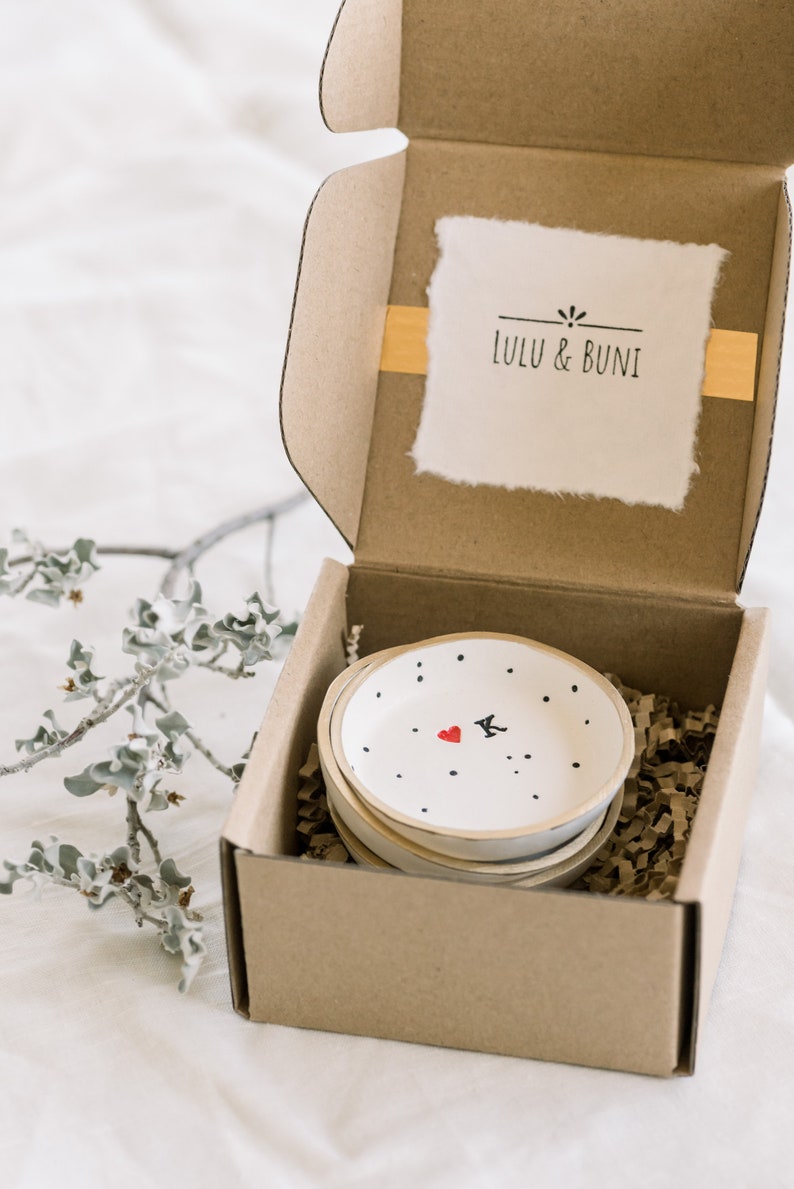 May include: A set of four small white ceramic dishes with black polka dots and a red heart with a black letter K in the center. The dishes are stacked inside a brown cardboard box with a white label that says "LULU & BUNi".