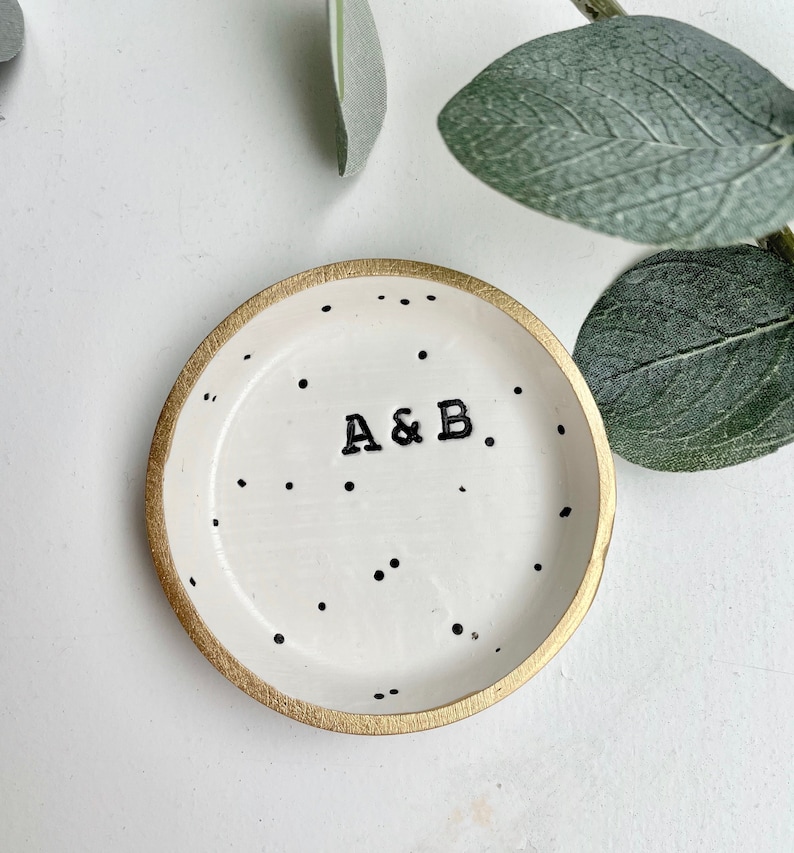 May include: A white ceramic ring dish with a gold rim and black polka dots. The dish has the initials "A&B" written in black.