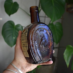 May include: A brown glass bottle shaped like a horseshoe with a horse head embossed in the centre and the text Horse Shoe Brand. A paper label on the side reads Grandam Heaton Receipt White Wine Vinegar with additional printed text.