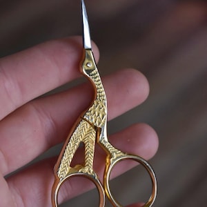 May include: Gold-colored embroidery scissors with a decorative bird-like design. The scissors have a sharp, pointed blade and round finger holes. The metal is etched with a textured pattern. A useful tool for sewing and crafting.