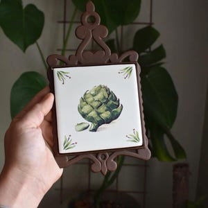 May include: A square ceramic tile featuring a painted green artichoke illustration, set within a dark brown decorative metal frame with an ornate top handle. The tile has a white background with small green and purple floral accents in the corners.