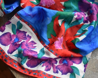 May include: A colorful silk scarf with a floral pattern. The scarf features large purple, blue, and red flowers against a white background. The edges are trimmed with a red border. The scarf is folded and draped.
