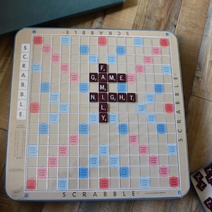 Pode incluir: Um jogo de tabuleiro Scrabble com as palavras "GAME NIGHT" escritas no centro. O tabuleiro é bege com quadrados vermelhos, azuis e rosa. O jogo está em uma superfície de madeira.
