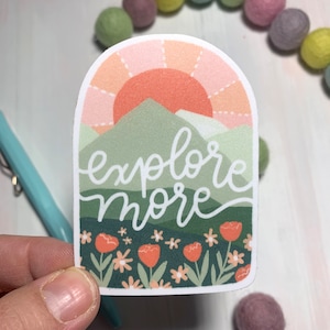 May include: A sticker with a sunset over mountains and the text "explore more" in a green and white font. The sticker has a pink, orange, and green color scheme with a field of flowers at the bottom.