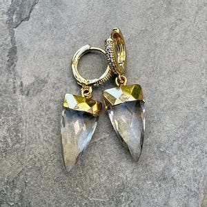 May include: A pair of gold-colored hoop earrings with clear, pointed crystal pendants. The earrings have a geometric design at the top of the crystal and are set against a gray background. The hoops are adorned with small, clear stones.