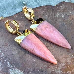 May include: A pair of gold hoop earrings with pink teardrop-shaped stones. The stones have a gold trim around the top.