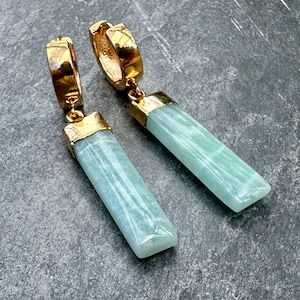 May include: A pair of gold-colored hoop earrings with rectangular, light blue-green gemstone pendants. The earrings have a gold cap at the top of the gemstone. The earrings are displayed on a dark gray surface.