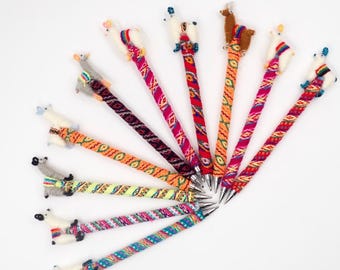 May include: A set of 12 colorful pens with llama toppers. The pens are wrapped in colorful patterned fabric and have silver tips. The llamas are made of wool and have different colored accents.