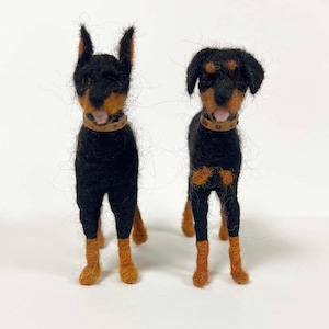 May include: Two handmade felt Doberman Pinscher dog figurines. The dogs are black with tan markings on their legs, chest, and face. They each wear a brown collar. One dog has upright ears, and the other has floppy ears.