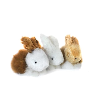 May include: Three plush toy bunnies in a row. One bunny has brown and white fur, the second is white, and the third is tan. The bunnies have small black eyes and long ears. The background is white.