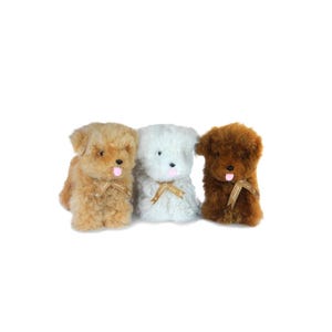 May include: Three plush toy puppies in shades of tan, white, and brown. Each puppy has a pink tongue, black eyes, and a ribbon around its neck. The toys are positioned against a white background.