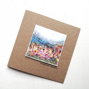 Può includere: Un biglietto con un dipinto ad acquerello di un campo di fiori selvatici. Il dipinto è incorniciato da un bordo di carta marrone e il testo "wildflower meadow" è stampato sotto il dipinto.