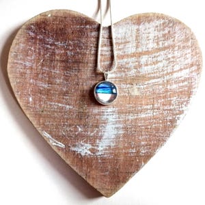 May include: A silver chain necklace with a round pendant featuring blue and white layered glass. The pendant is displayed on a heart-shaped wooden surface.
