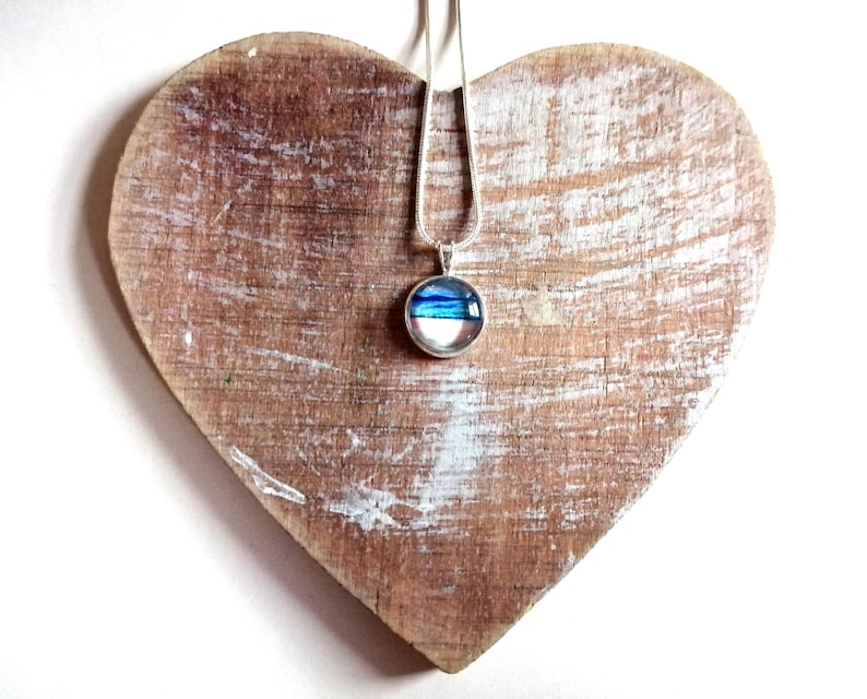 May include: A silver chain necklace with a round pendant featuring blue and white layered glass. The pendant is displayed on a heart-shaped wooden surface.