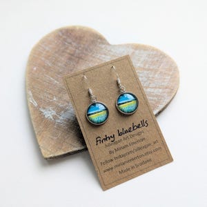 Può includere: Orecchini pendenti con design rotondo blu, giallo e verde, in metallo argentato. Sono presentati su una scheda marrone con il testo "Fintry bluebells". La scheda poggia su un oggetto di legno a forma di cuore.