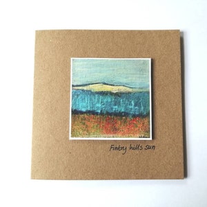 Può includere: Un biglietto marrone con un piccolo dipinto di un paesaggio blu e verde con una collina bianca sullo sfondo. Il testo "Fintry hills sun" è scritto sotto il dipinto.