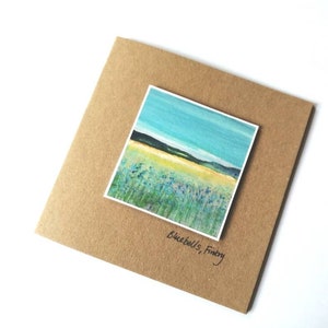 Può includere: Un biglietto marrone con un piccolo dipinto di un cielo blu, erba verde e fiori gialli. Il dipinto è intitolato "Bluebells, Fintry".