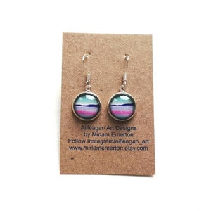 May include: A pair of silver dangle earrings with round, colorful, layered glass cabochons. The cabochons feature a pink, purple, blue, and green color scheme. The earrings are on a brown card with the text "Ailleagan Art Designs by Miriam Emerton Follow Instagram/ailleagan_art www.miramemerton.etsy.com"