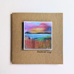 Può includere: Un piccolo dipinto quadrato di un tramonto su una baia, con colori vivaci di rosa, arancione e blu. Il dipinto è montato su una carta marrone quadrata più grande. Il testo "Sheltered bay" è scritto sotto.
