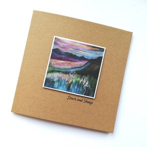 Può includere: Un biglietto marrone con un dipinto di paesaggio colorato di una catena montuosa con un cielo rosa e viola. Il dipinto è intitolato "Stars and Sheep".