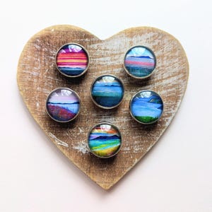 May include: Six round, silver-rimmed landscape art magnets are arranged on a heart-shaped wooden board. The miniature paintings depict colorful sunsets, mountains, and bodies of water. The wooden board has a natural, weathered appearance.