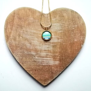 Può includere: Una collana color oro con un pendente rotondo con un design astratto blu e verde. Il pendente è appeso a una catena sottile. La collana è esposta su una superficie di legno a forma di cuore.