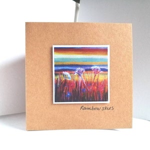 Può includere: Biglietto di auguri quadrato marrone con un dipinto colorato di un campo e un cielo arcobaleno. Il dipinto è in una cornice bianca. Le parole "Rainbow skies" sono scritte in corsivo in basso sul biglietto.