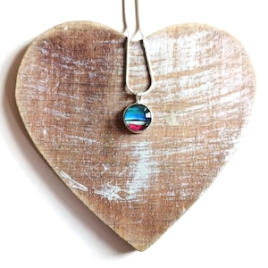 May include: A silver chain necklace with a round pendant featuring a colorful abstract design. The pendant is on a wooden heart-shaped surface.