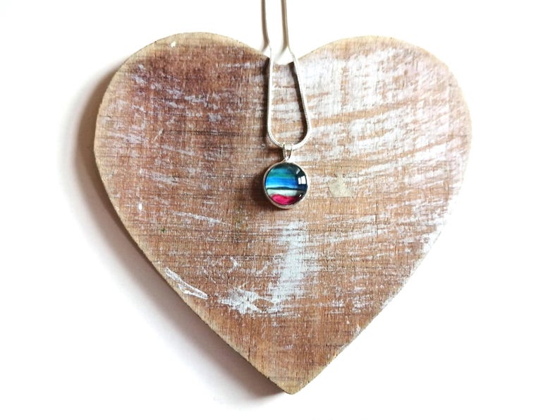 May include: A silver chain necklace with a round pendant featuring a colorful abstract design. The pendant is on a wooden heart-shaped surface.