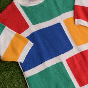Puede incluir: Un suéter colorido con un diseño geométrico. El suéter presenta grandes bloques de color rojo, verde, azul y amarillo, separados por rayas blancas. Las mangas y el cuello están acanalados con ribetes blancos. El suéter está hecho de un material de punto suave.