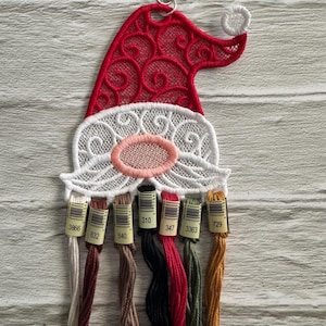 May include: A red and white Santa hat-shaped thread organizer with a silver clasp. The hat features intricate lace-like detailing. Below the beard are several spools of thread in various colors, including beige, brown, red, and green.