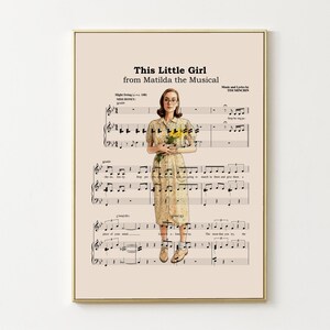 May include: A framed print featuring sheet music with the title "This Little Girl from Matilda the Musical." The image includes a woman holding flowers, set against a beige background. The print has a gold frame.