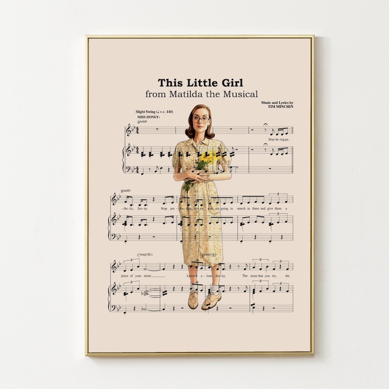 May include: A framed print featuring sheet music with the title "This Little Girl from Matilda the Musical." The image includes a woman holding flowers, set against a beige background. The print has a gold frame.