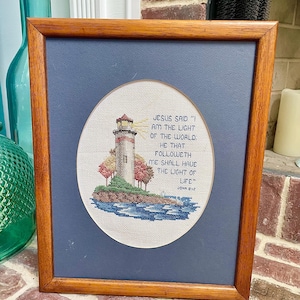 May include: A framed cross-stitch embroidery featuring a lighthouse on a small island surrounded by blue water. The text reads: "JESUS SAID 'I AM THE LIGHT OF THE WORLD: HE THAT FOLLOWETH ME SHALL HAVE THE LIGHT OF LIFE' JOHN 8:12". The frame is wood.