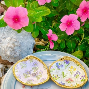 May include: Two decorative seashell-shaped ring dishes with gold trim, one with a floral pattern and the other with a lavender and green pattern. A ring rests in one dish. Pink flowers and green leaves are in the background.