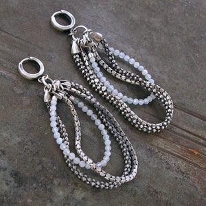 May include: A pair of silver drop earrings. Each earring has a silver hoop and multiple strands of silver chain and small, pale blue beads. The chains are looped and connected at the top, creating a layered effect. The earrings are displayed on a textured, dark surface.