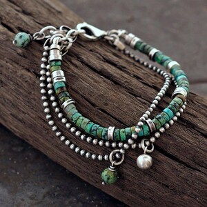 Op de afbeelding: Een armband met meerdere strengen, voorzien van turquoise heishi-kralen, zilveren kogelketting-schakels en gladde zilveren cilindrische tussenstukjes. De armband bevat twee bungelende bedels: een ronde zilveren kraal en een ronde turquoise kraal.