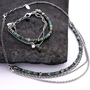 May include: A multi-strand necklace and bracelet set featuring small, cylindrical turquoise beads interspersed with silver-toned metal spacers. The jewelry includes delicate silver chains, a lobster clasp, and small dangling charms, including a single white bead.