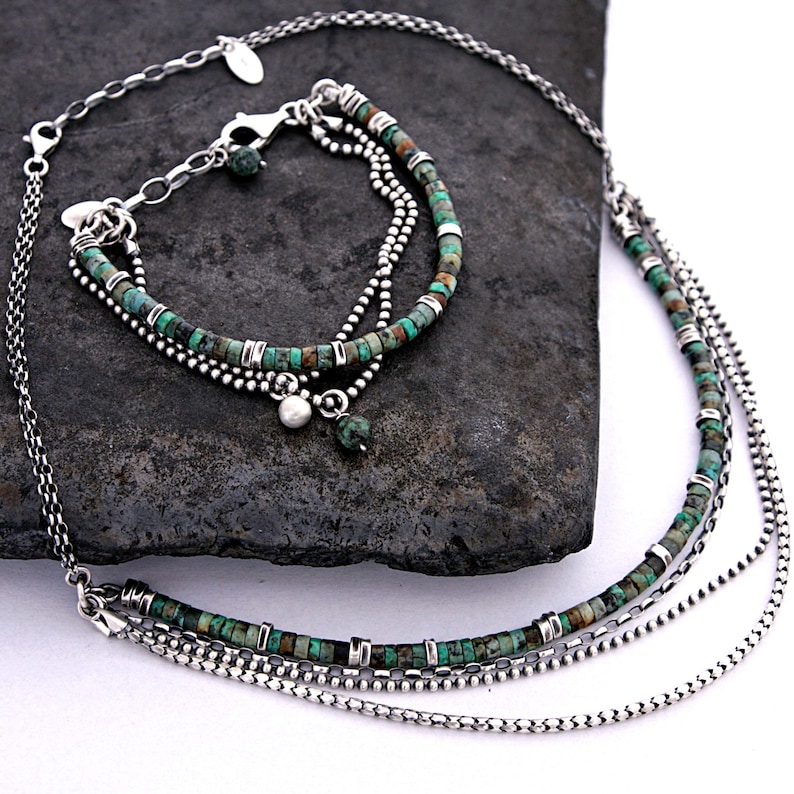 May include: A multi-strand necklace and bracelet set featuring small, cylindrical turquoise beads interspersed with silver-toned metal spacers. The jewelry includes delicate silver chains, a lobster clasp, and small dangling charms, including a single white bead.