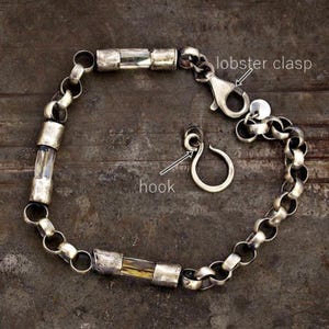 May include: Silver bracelet with a lobster clasp and a separate hook. The bracelet has a chain link design with cylindrical, clear and amber-coloured accents. The bracelet is displayed on a dark, textured surface.