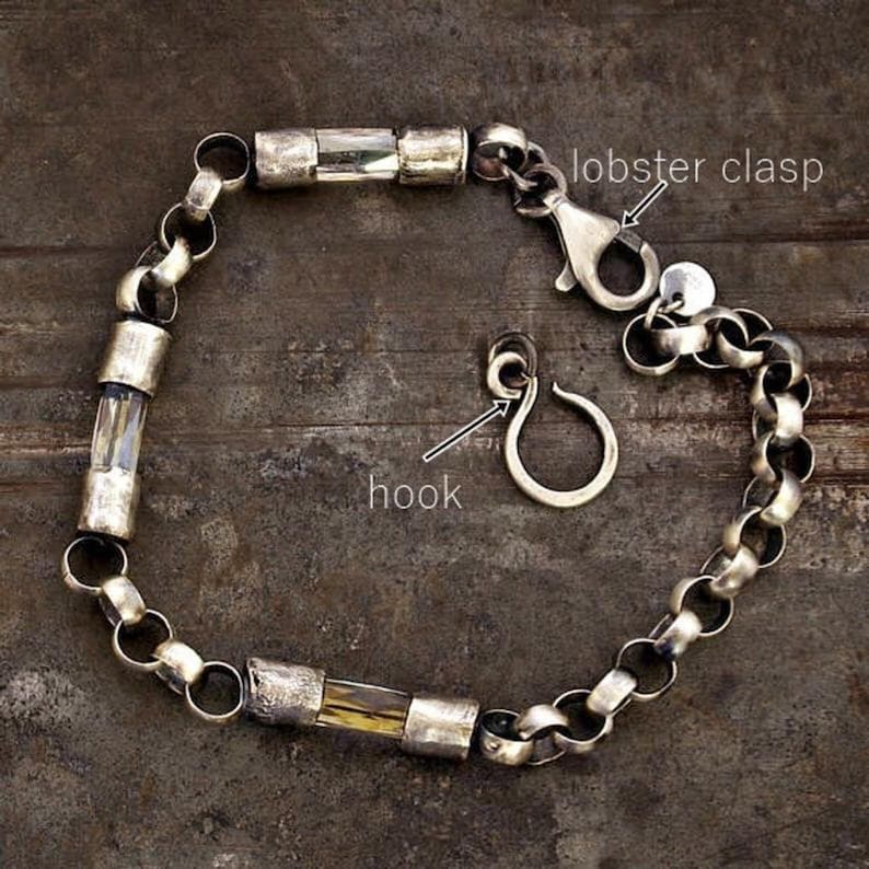 May include: Silver bracelet with a lobster clasp and a separate hook. The bracelet has a chain link design with cylindrical, clear and amber-coloured accents. The bracelet is displayed on a dark, textured surface.